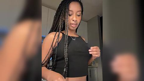 Media: Video of a young Black woman with braided hair in a black crop top, standing in a modern kitchen, looking down.