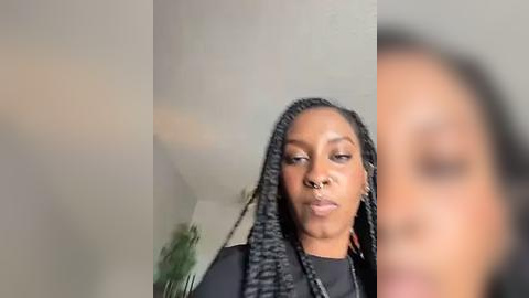 Media: A video of a Black woman with braided hair, wearing a black jacket, standing in front of a gray wall with blurred greenery in the background.