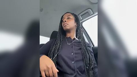 Media: Video of a young Black woman with long braids, wearing a dark blue cardigan, sitting in a car, looking introspective. The car's interior is dimly lit, with blurred window reflections in the foreground.