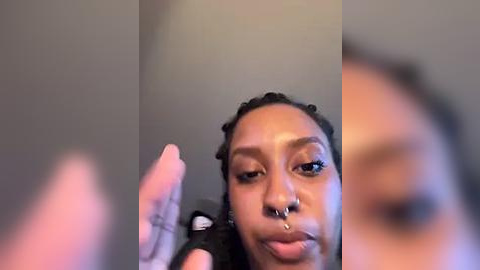 Media: Video of a Black woman with a medium-brown complexion, wearing a nose ring and large hoop earrings, adjusting her hair in a mirror. Background is blurred, focusing on her face.