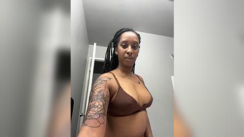 Media: Video of a curvy Black woman with medium brown skin, long braids, and a tattooed arm in a brown bra, standing indoors against a white wall.