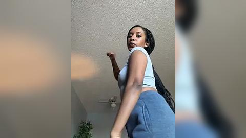 Media: Video of a young Black woman with medium skin tone, wearing a white crop top and blue shorts, mid-stride, indoors with a textured ceiling and a blurred background.