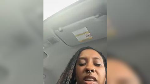 Media: Video of a young Black woman with medium-brown skin and long, curly hair, wearing a nose ring, looking up from a seated position in an airplane. The image is slightly out of focus, capturing the cabin ceiling with a safety card visible.