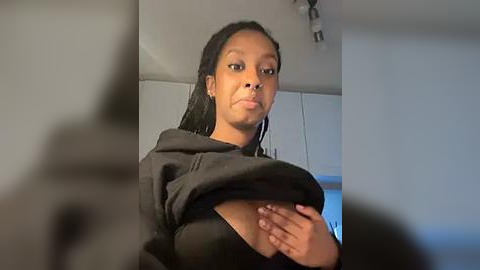 Media: Video of a young Black woman with medium-brown skin, pulling up her black shirt to reveal her small, dark areolas and nipples. She has long, straight hair. Background shows a kitchen with white cabinets and a ceiling light.