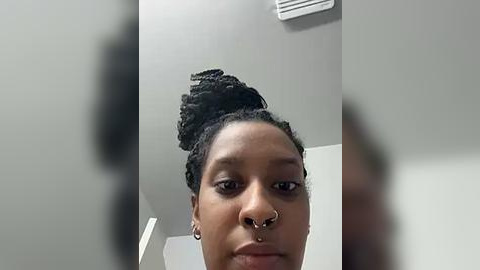 Media: Video of a Black woman with dark skin, short curly hair in a bun, and a septum piercing, standing in a white room with an air conditioner above her.