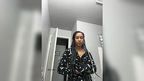 Media: Video of a young woman with medium-brown skin and straightened black hair, wearing a black robe with white floral patterns, standing in a modern, minimalist bathroom with white walls and a silver towel rack.