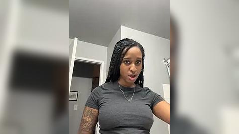 Media: A video of a Black woman with braided hair, wearing a grey t-shirt, standing indoors near a door. She has a tattoo on her left arm. The room is minimally decorated with white walls and a framed picture.