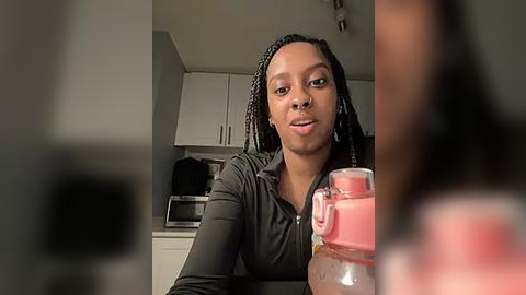 Media: Video of a Black woman with braided hair, wearing a black shirt, holding a pink drink in a glass container, sitting in a modern kitchen with white cabinets and stainless steel appliances.