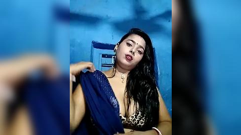 Media: Video of a Latina woman with long black hair, medium skin tone, and a voluptuous figure, wearing a leopard print top, holding a blue garment in a dimly lit room with blue walls.