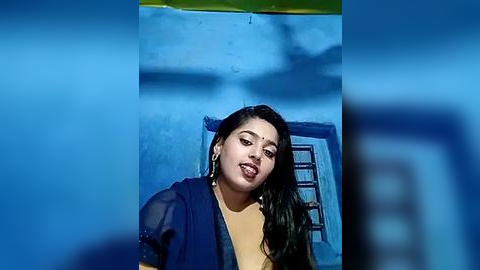 Media: Video of a young woman with long, dark hair, wearing a blue shawl over a tan top, against a blue-painted wall with a window, capturing a casual, candid moment.