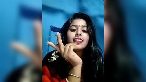 Media: Video of a young South Asian woman with long black hair, wearing a red and black traditional dress, making a peace sign. Background is blurred blue.