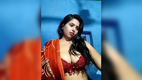 Media: Video of a curvy South Asian woman with long black hair, wearing a red lace bra and orange sari, against a blue background.