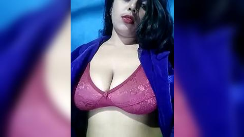 Media: Video of a curvy woman with long black hair, wearing a red lace bra and blue jacket, against a blurred blue background.