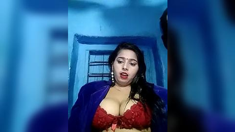 Media: Video of a woman with long black hair, fair skin, and large breasts wearing a red lace bra and purple jacket, standing in a dimly lit blue room with a barred window.