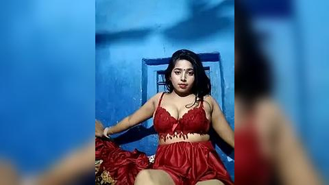 Media: A video of a young woman with medium skin tone and long black hair, wearing a red lace lingerie set, seated on a blue chair in a dimly lit room with blue walls.