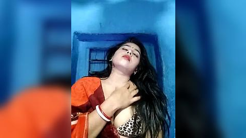 Media: Video of a woman in an orange sari, eyes closed, leaning against a blue wall, wearing a leopard-print bra, hands touching her chest, indoors.