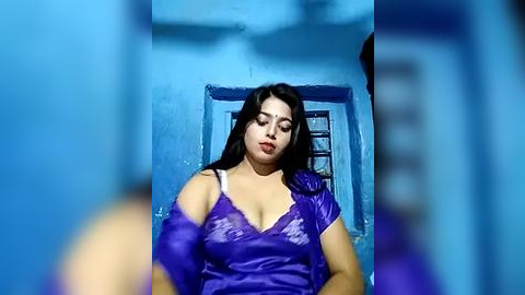 Media: A video of a young South Asian woman with medium skin tone, wearing a purple blouse and standing in a blue-painted room with a window.