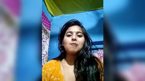 Media: Video of a young South Asian woman with long black hair, wearing a mustard-yellow blouse, standing under a blue umbrella in a colorful, sunlit outdoor setting.