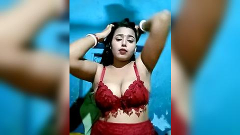 Media: Video of a Latina woman with medium skin tone, wearing red lace lingerie, sitting on a chair against a blue wall, with blurred figures in the background.