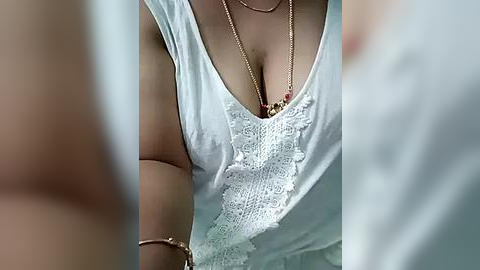 Media: Video of a woman's torso in a white lace top, with delicate floral embroidery. She wears a gold necklace with a pendant and a bracelet. The background is blurred, focusing on the intricate details of her top.