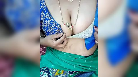 Media: Video of a woman in a blue and white traditional dress, revealing her cleavage as she adjusts her bra, with a green sari partially covering her abdomen.