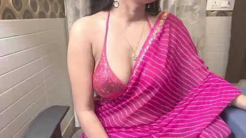 Media: Video of a South Asian woman with medium skin tone, wearing a pink sequined bra and a pink and white striped sari, sitting on a medical examination table in a clinic with beige and white tiled walls.