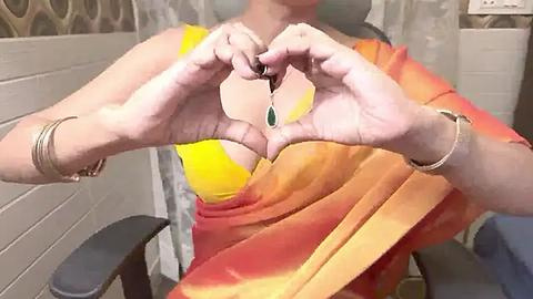 Media: Video of an Indian woman with light skin and medium build, wearing a vibrant yellow and orange sari, forming a heart with her hands, showcasing a green pendant necklace. Background includes beige tiled walls and a black chair.