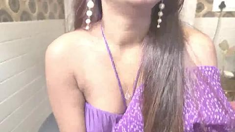 Media: Video of a woman with long, straight brown hair wearing a purple off-the-shoulder top and dangling pearl earrings, set against a tiled wall with circular patterns.