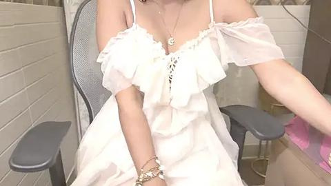 Media: A video of a woman in an off-shoulder white dress with ruffled sleeves, sitting on a gray mesh chair. She wears a gold necklace and bracelet, against a beige brick wall background.