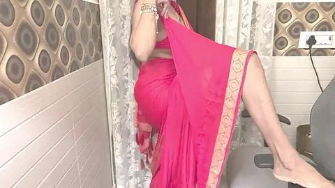 Media: Video of a woman in a bright pink sari, sitting on a grey chair, wearing a bracelet, with a textured wall and patterned curtain in the background.