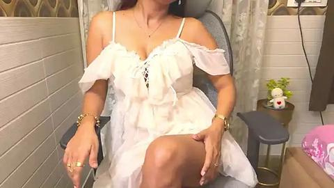 Media: Video of an Asian woman in a white off-shoulder dress, seated on a grey chair in a room with floral wallpaper, a plant, and a pink teddy bear.