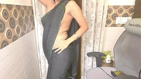 Media: Video of a light-skinned, slender woman with a small chest, wearing a black sleeveless dress with a side slit, posing in a modern room with white and brown patterned wallpaper and a gray gaming chair.