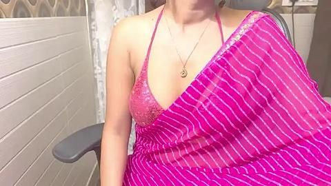 Media: Video of a light-skinned woman wearing a pink, sequined halter top, draped in a pink striped sari, seated on a gray chair against white and beige tiled walls in a modern bathroom.