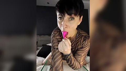 Media: Video of a woman with short dark hair, wearing a striped shirt, licking a pink lollipop, sitting on a bed in a dimly lit bedroom.