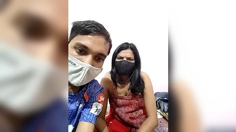 Media: Video of a man with a blue shirt and face mask sitting next to a woman in a red dress, both with black masks, in a dimly lit room.