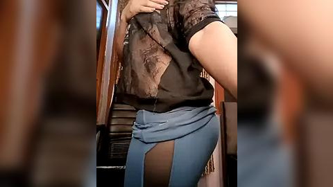 Media: Video of a person with light skin, wearing a black lace top and blue jeans with a slit revealing a black thong. The background shows a blurred indoor setting with wooden furniture.