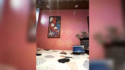 Media: Video of a cozy bedroom with pink walls adorned with abstract floral decals, featuring a laptop on a bed with a polka-dot comforter. A framed artwork with vibrant colors hangs on the wall.