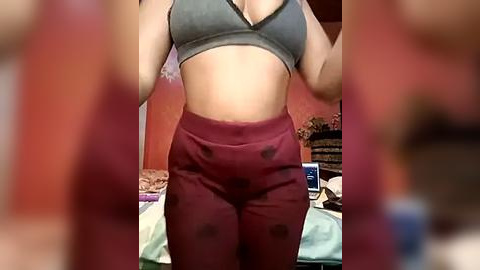 Media: Video of a woman with light brown skin wearing a gray sports bra and maroon leggings, standing in a cluttered bedroom with a bed, a laptop, and a wall-mounted TV.