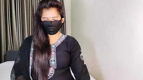 Media: Video of an Asian woman with long black hair, wearing a black face mask, black long-sleeved top with geometric patterns, and holding a black bag, standing indoors with grey curtains in the background.