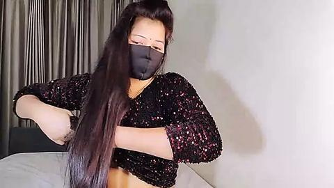 Media: Video of an Asian woman with long, straight black hair, wearing a black sequined top, black face mask, and a red bindi, standing against a gray curtain backdrop, arms crossed.