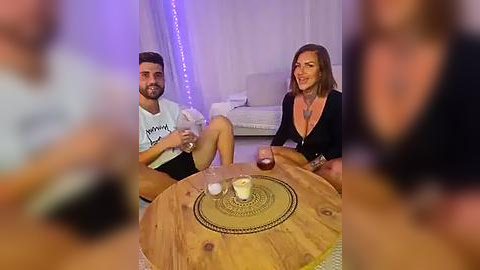 Media: Video of a couple sitting on a wooden round table with a decorative mat, drinking water, in a dimly lit room with white walls and soft purple lights.