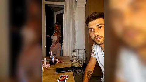 Media: Video of a man with a tattooed arm, sitting at a wooden table with cards, wine glasses, and a phone, while a woman in a white dress stands in the background.