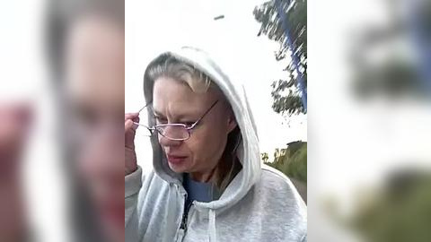 Media: Video of a middle-aged woman with glasses and blonde hair, wearing a light gray hoodie, adjusting her glasses outdoors. The background features blurred greenery and a tree.