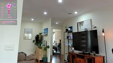 Media: Video of a modern, brightly lit living room with a large flat-screen TV, wooden entertainment center, plants, and abstract art on beige walls.