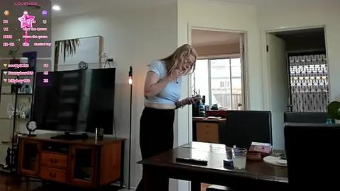 Media: Video of a blonde woman in a light blue shirt and black pants, standing in a modern living room with a flat-screen TV, wooden furniture, and a computer desk.