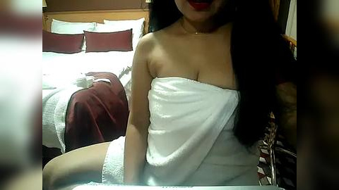 Media: Video of an Asian woman with long black hair, wearing a white towel, sitting on a bed with red and white bedding.