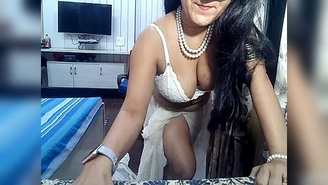 Media: A video of a smiling, light-skinned woman with long black hair, wearing a white lace bra and sheer dress, sitting on a bed with a blue patterned sheet. She has a pearl necklace and a smartwatch on her wrist. The room has white paneling, a TV, and a dresser in the background.