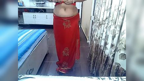 Media: A video of a woman with a light brown complexion, wearing a red saree with gold floral patterns, standing in a modestly furnished bedroom.