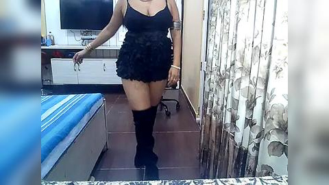 Media: A video of a woman with medium skin tone, wearing a black mini-dress and thigh-high boots, standing in a bedroom with a TV, patterned curtains, and a bed.