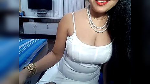 Media: Video of a smiling woman with medium-dark skin, wearing a white dress, pearls, and a gold bracelet, sitting on a bed with blue sheets in a modern bedroom.